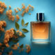 A clear glass perfume bottle with amber liquid and a dark cap sits on a blue surface next to a branch of small orange flowers and green leaves.
